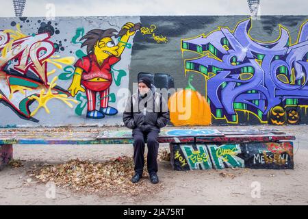 Älterer Mann, der auf einer Bank neben der Street Art an der ehemaligen Berliner Mauer im Mauerpark, Prenzlauer Berg, Berlin, sitzt Stockfoto