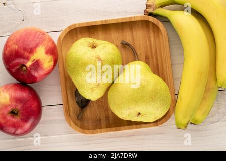 Zwei saftige Bio-Birnen, ein Bund Bananen und zwei rote Äpfel mit einem Bambustablett auf einem Holztisch, Nahaufnahme, Draufsicht. Stockfoto