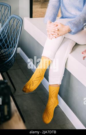 Tröstende, gemütliche Frau in gestrickten woll-Wintersocken, weißen Hosen und blauem warmen Pullover, die im Winter auf einer Bank im Café sitzen Stockfoto