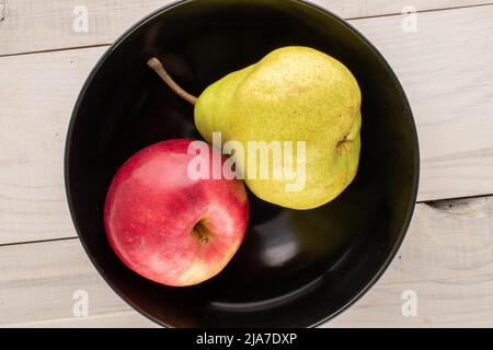 Eine saftige Bio-Birne und ein roter Apfel in einer schwarzen Keramikplatte auf einem Holztisch, Nahaufnahme, Draufsicht. Stockfoto