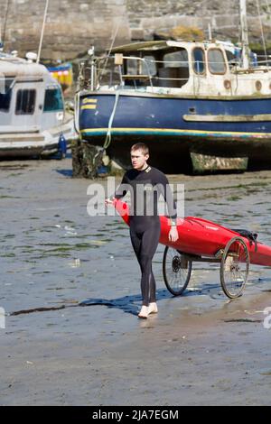 Junger Mann im Neoprenanzug mit seinem Kajak auf einem Trolley im Hafen von Porthleven bei Ebbe Stockfoto