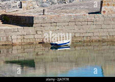 Leeres blau-weißes Ruderboot vor der Hafenmauer aus Granit in Porthleven, Cornwall. Das Ruderboot spiegelt sich in den ruhigen Gewässern. Stockfoto