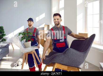 Zwei glückliche junge Arbeiter in einem umzugsenden Unternehmen, die Stühle, Sessel und Pflanzen tragen Stockfoto