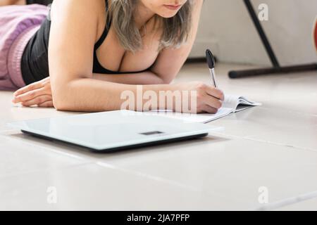 Nahaufnahme Detail einer Latina-Frau, auf dem Boden liegend, sehr nachdenklich, Schreiben auf ihr Notizbuch die Ziele und Ziele ihrer täglichen Ernährung. Mädchen Kalkül Stockfoto