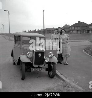 1950s, im historischen Großbritannien der Nachkriegszeit und auf einem Bürgersteig in einer Wohnsiedlung, stehen ein junger Mann, seine Freundin und Mutter zusammen für ein Foto von ihrem Auto, einer Austin Seven. Der britische Made Austin 7 war ein kleines - mit dem Spitznamen "Baby Austin" - Economy-Auto, das von 1923 bis 1939 produziert wurde und das Fahrzeugeigentum an diejenigen brachte, die es sich nie zuvor leisten konnten, und beworben als: "Ein Penny-a-Mile mit vier mehr deckt die laufenden Kosten". Es wurde auch Frauen als „das Auto für die weibliche Note“ beworben. Der Wagen war in Großbritannien bis in den 1960s noch im Einsatz. Stockfoto