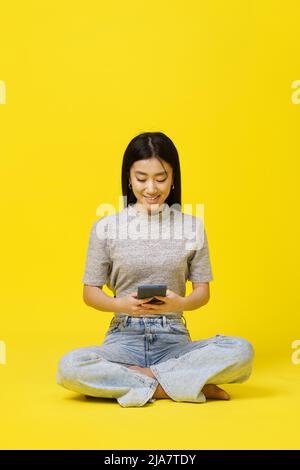 Asiatische junge Mädchen auf dem Boden sitzend Smartphone SMS oder Online-Shopping isoliert auf gelbem Hintergrund. Produktplatzierung für mobile App-Werbung. Speicherplatz kopieren. Stockfoto