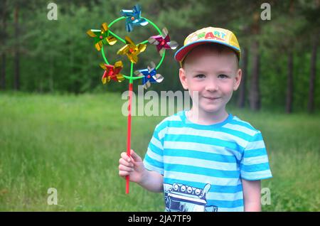 Glückliche Kindheit - Konzept. Der kaukasische Junge lächelt in einer Mütze und einem gestreiften T-Shirt mit einer Windmühle in der Hand vor dem Hintergrund eines grünen Waldes. Stockfoto
