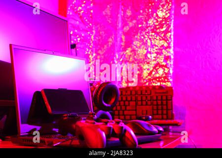 Arbeitsbereich des Spielers. Gamepads, Computermaus, Kopfhörer, Tastatur und Laptop auf dem Tisch. Gaming-Zubehör und Computer-Ausrüstung in Neonlicht. Ga Stockfoto