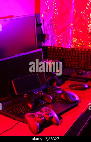 Arbeitsbereich des Spielers. Gamepads, Computermaus, Kopfhörer, Tastatur und Laptop auf dem Tisch. Gaming-Zubehör und Computer-Ausrüstung in Neonlicht. Ga Stockfoto