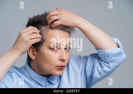 Portrait einer reifen Frau im Jahr 50s, die ihre Haare überprüft und unglücklich war, ein Ergebnis im Spiegel zu sehen. Schöne grauhaarige Frau, die mit Schuppen zu tun hat, Problem beim Einchecken im Spiegel. Stockfoto