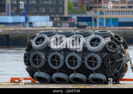 Ein großer Gummiboot-Stoßfänger am Rand eines Docks. Oberfläche mit alten Reifen bedeckt, die von Ketten gehalten werden.Wasser und Gebäude im Hintergrund. Stockfoto