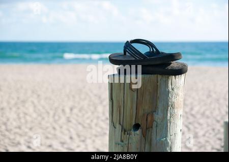 Ein Paar Flip-Flops, die auf einem verwitterten Mast am Strand vor einer Horizont-Kulisse aus Sand, Meerwasser und Himmel zurückgelassen wurden Stockfoto