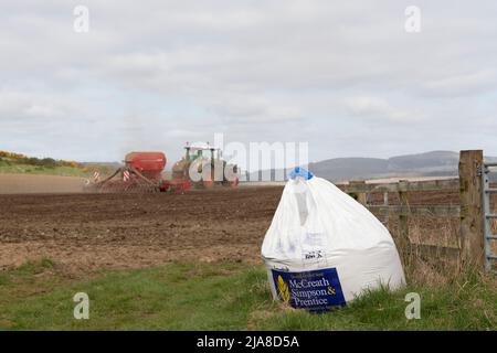 Ein Sack des Händlers mit Saatgut, der neben dem Eingang zu einem gepflügten Feld sitzt, mit einem grünen Traktor, der eine Sämaschine schleppt, die im Hintergrund in Betrieb ist Stockfoto