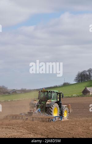 Ein Landwirt mit einem grünen John Deere Traktor und einem Scarifier, der in Spring Sunshine ein gepflügeltes Feld ausfüllt Stockfoto