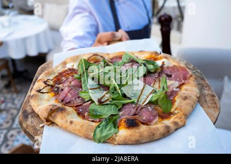 Pizzaplatte aus Holz in den Händen des Küchenchefs und Fleischpizza mit Mozzarella-Käse und Rucola-Blättern Stockfoto