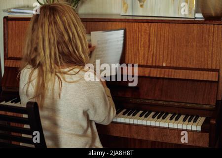 Rückansicht einer Musikerin mit welligem, hellem Haar, die am alten braunen Klavier sitzt und Notenblätter mit Musiknoten hält. Stockfoto