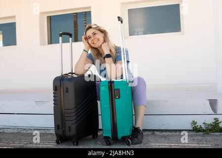 Blonde lächelnde Frau sitzt auf dem Boden des Bahnhofs und lehnt sich an das Gepäck. Reise oder Reise starten. Vorfreude auf Neues Stockfoto