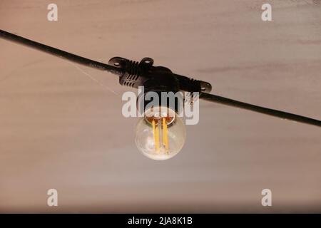 Eine runde, energieeffiziente LED-Glühlampe aus klarem Glas, die unter der Decke eines Terrassendachs des Restaurants hängt Stockfoto