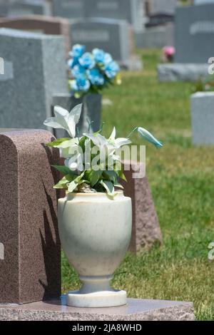 Granitvase aus künstlichen Allwetter-Stofflilien blüht neben Grabstein auf dem Friedhof Stockfoto