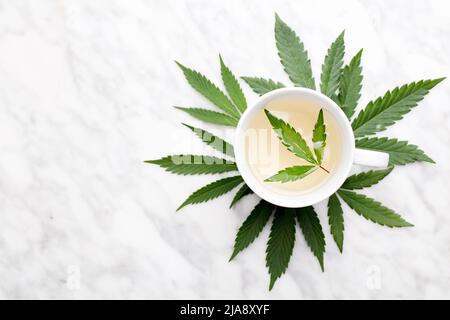 Weißer Becher mit Marihuana-Infusion mit grünem Blatt innen auf Marmortisch mit Blättern. Draufsicht. Stockfoto