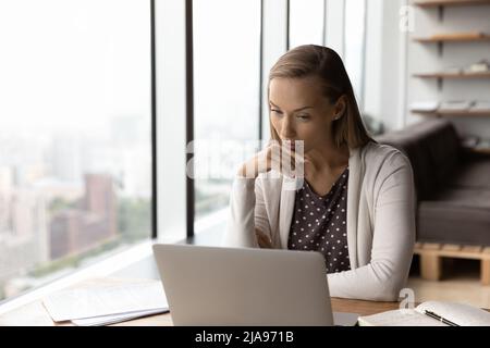 Durchdachte Geschäftsfrau, die am Laptop arbeitet und Text auf dem Bildschirm liest Stockfoto