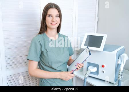 Der Kosmetologe steht an der Laser-Haarentfernungsmaschine mit Ordner für die Aufzeichnung von Kunden und lächelt. Frau im grünen Mantel Arzt des Schönheitssalons wartet auf Kunden Stockfoto