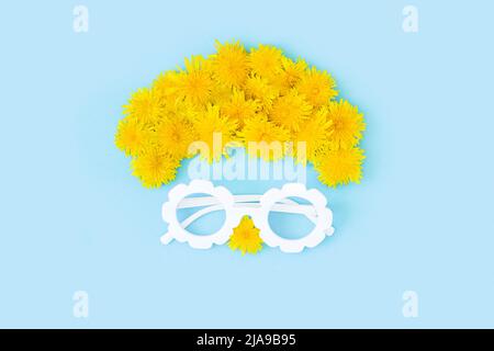 Kreatives Sommerkonzept. Abstraktes Gesicht mit Brille und gelben Delelions-Blüten auf blauem Hintergrund. Draufsicht Flat Lay. Stockfoto