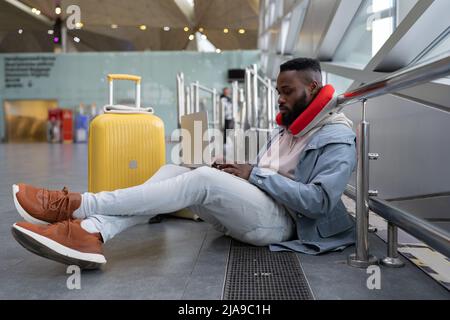 Nachdenklicher junger schwarzer Mann sitzt mit gekreuzten Beinen zusammen und wartet auf den Flug mit einem Computer neben dem Fenster. Stockfoto