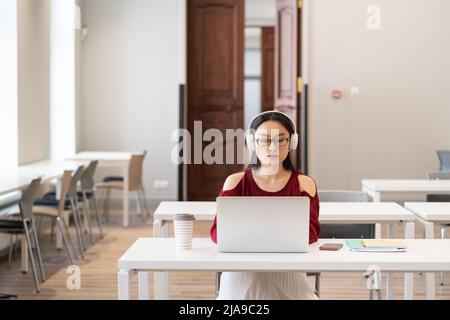 Serious Asian Student Mädchen in Kopfhörern beobachten pädagogische via Internet auf Laptop in der Bibliothek Stockfoto