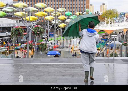 Fußgänger & Regenschirm Frühling April Duschen regnerischer Tag Spaziergang Westfield Fußgängerbrücke in Richtung Shoal Titanium Fisch Skulpturen Stratford London England Großbritannien Stockfoto