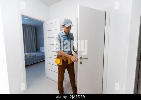 Installation eines Schlosses an der vorderen hölzernen Eingangstür. Porträt eines jungen Schlosserarbeiters in blauer Uniform, der Türknauf installiert. Professionell Stockfoto