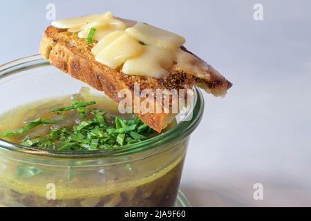 Zwiebelsuppe mit frischen grünen Kräutern im Glas und geröstetes Brot mit geschmolzenem Käse auf grauem Hintergrund, Kopierraum, ausgewählter Fokus, schmal Stockfoto