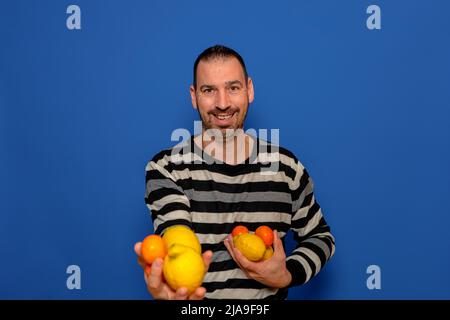 Bärtiger hispanischer Mann, der einen gestreiften Pullover trägt, bietet aufgeregt Orangen und Zitronen an, die er isoliert im blauen Studio in seinen Händen hält Stockfoto