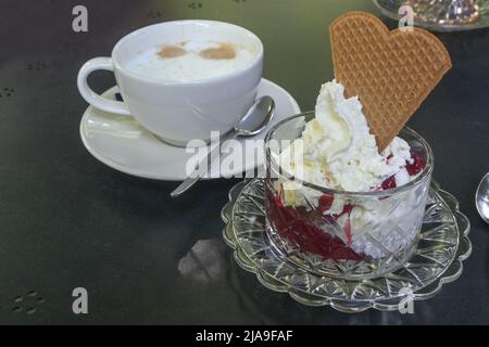 Eis mit roten Früchten und Schlagsahne in einer Glasschüssel und eine Tasse Kaffee auf einem dunklen Tisch in einem Straßencafé, Kopierraum, ausgewählter Fokus, schmal d Stockfoto