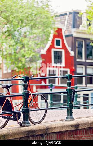 Orangefarbenes holländisches Fahrrad, das an Geländer über einen Kanal in Amsterdam mit einem roten traditionellen niederländischen Haus angekettet ist Stockfoto