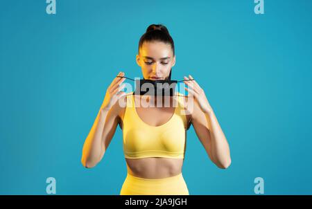 Fitness-Frau, Die Eine Chirurgische Gesichtsmaske Auf Blauem Hintergrund Aufsetzt Stockfoto