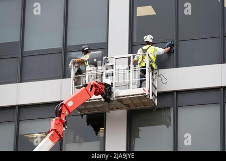 Männer, die in einem mehrstöckigen Büro in Basingstoke in einer mobilen erhöhten Arbeitsbühne (MEWP) mit persönlicher PSA und Reinigungsfenstern arbeiten. England Stockfoto