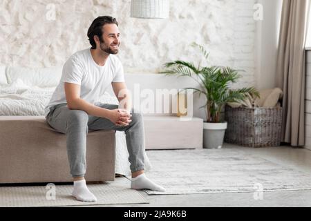 Glücklicher bärtiger junger Mann, der zu Hause auf dem Bett sitzt Stockfoto