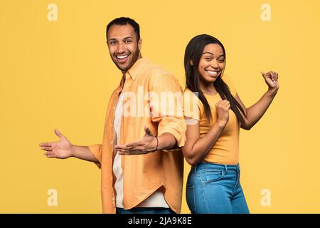 Fröhliches junges schwarzes Paar tanzt, feiert gemeinsam Erfolg auf gelbem Studiohintergrund Stockfoto