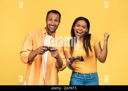 Aufgeregt junge afroamerikanische Paar mit Joysticks spielen Computerspiele, gestiking JA auf gelben Studio-Hintergrund Stockfoto