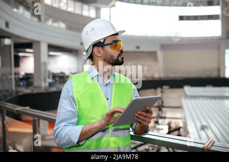 Serious nachdenklich tausendjährigen islamischen männlichen Ingenieur in Schutzuniform, Brille und Hut mit Bart mit Tablette Stockfoto