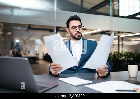 Konzentrierter, vielbeschäftigter, gutaussehender, tausendjähriger islamischer Geschäftsmann mit Bart in einer Brille arbeitet mit Laptop und Dokumenten Stockfoto