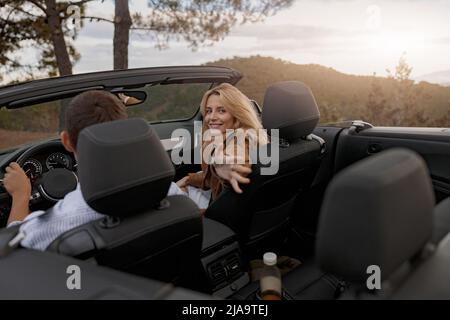 Fröhliche kaukasische junge hübsche Frau, die im Cabrio auf dem Beifahrersitz sitzt und ihre Hand heraushält Stockfoto