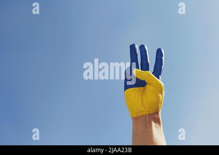 Die sonnenbeschienene Hand des Mannes, gemalt in Farben der ukrainischen Nationalflagge, zeigt eine traditionelle dreizackige Handbewegung als Symbol für Unterstützung und Solidarität Stockfoto