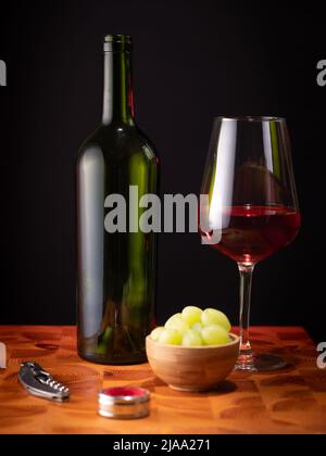 Helsinki / Finnland - 29. MAI 2022: Eine leere Rotweinflasche, ein Glas Wein und einige Trauben auf einer Holzfläche vor dunklem Hintergrund. Stockfoto
