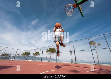 Vlore, ALBANIEN - 20. MAI 2022: Männlicher Basketballspieler schießt einen Sprung im Freien. Allein Ball spielen. Sonnenaufgang Ozean Hintergrund. Stockfoto