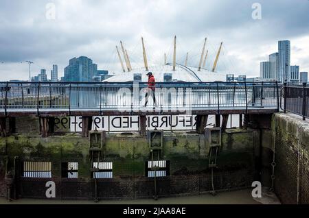 Der Mensch geht über den Beckensteg des East India Dock mit der Themse und der O2 Arena im Hintergrund. Stockfoto