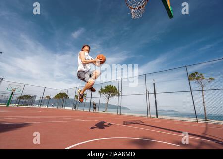 Vlore, ALBANIEN - 20. MAI 2022: Männlicher Basketballspieler schießt einen Sprung im Freien. Allein Ball spielen. Sonnenaufgang Ozean Hintergrund. Stockfoto