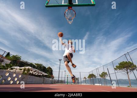 Vlore, ALBANIEN - 20. MAI 2022: Männlicher Basketballspieler schießt einen Sprung im Freien. Allein Ball spielen. Sonnenaufgang Ozean Hintergrund. Stockfoto