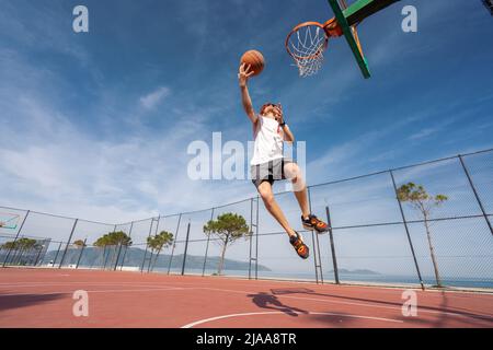 Vlore, ALBANIEN - 20. MAI 2022: Männlicher Basketballspieler schießt einen Sprung im Freien. Allein Ball spielen. Sonnenaufgang Ozean Hintergrund. Stockfoto